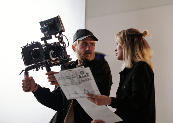 Young bearded cameraman and his assistant with documents consulting about shooting while standing in front of camera in studio